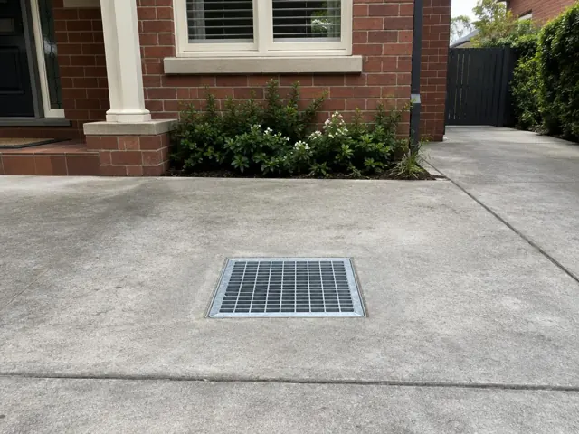 Clean outdoor drain grate in a concrete driveway beside a brick house in Gordon